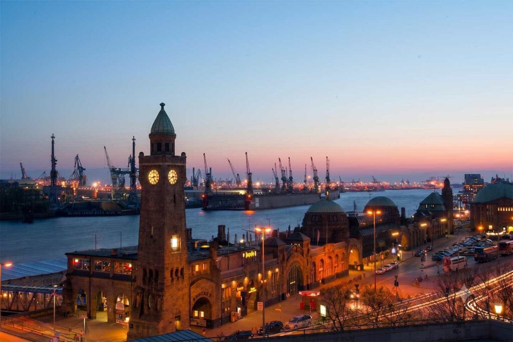 Abendaufnahme der beleuchteten Hamburger Landungsbrücken mit dem Pegelturm, Barkassen auf der Elbe und Containerfrachtern vor den Hafenkränen bei Dämmerung.